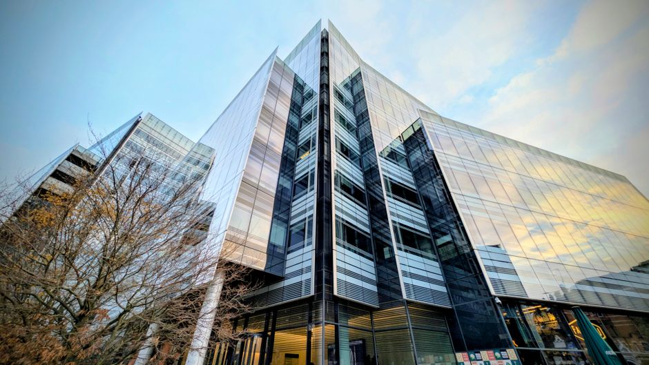 A modern glass office building at Chiswick Business Park, viewed from ground level looking upward, showcasing reflective glass panels and metal framing with a bright yellow sky in the background. The 