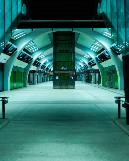 A well-lit indoor area, possibly part of a commercial or public space, featuring polished dark tiled flooring that reflects light. In the background, there is a staircase with stainless steel handrail